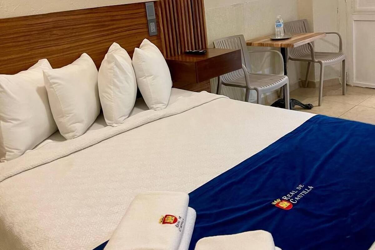 A hotel room with a neatly made bed, a desk with a chair, and a wall-mounted lamp.