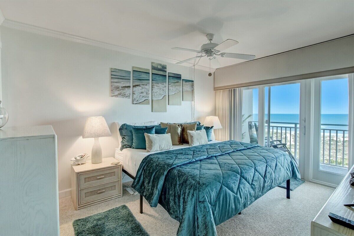 A bedroom with a bed, nightstands, a ceiling fan, and a view of the ocean through large windows.
