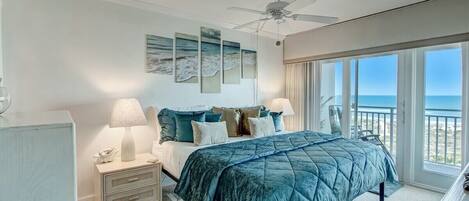 A bedroom with a bed, nightstands, a ceiling fan, and a view of the ocean through large windows.