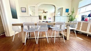 A dining area with a wooden table and four white wireframe chairs. There is a ceiling fan above the table, and a kitchen area with cabinets and appliances is visible in the background.