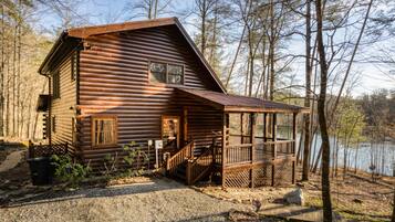 A rustic log cabin with a porch, surrounded by trees and a lake.