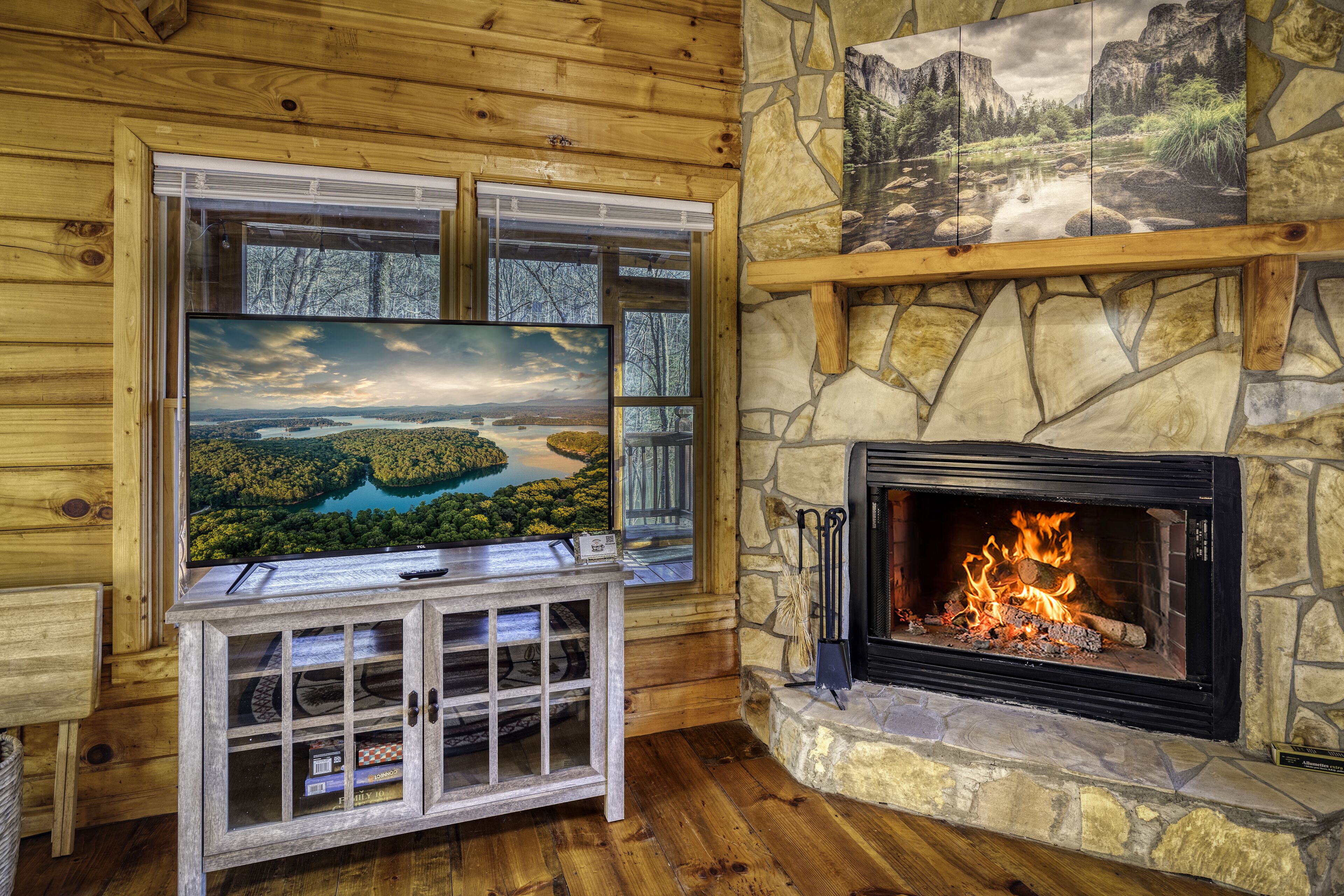 A cozy room with a stone fireplace, a large TV mounted on a wooden cabinet, and a scenic painting above the fireplace.