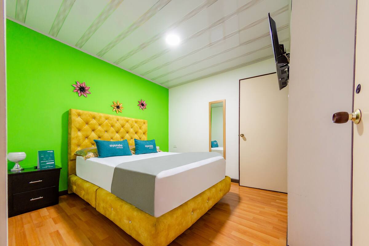 Een kamer met een bed met een geel bekleden hoofdeinde, groene wanden, een houten vloer, een wandtelevisie en een deur met een messing knop.