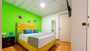 Una habitación con una cama de cabecera amarilla y tapizada, paredes verdes, piso de madera, televisor de pantalla plana montado en la pared y una puerta con manija de bronce.
