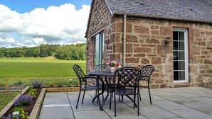 Un edificio de piedra con chimenea, un patio con sillas y una mesa, y vistas a un campo verde.