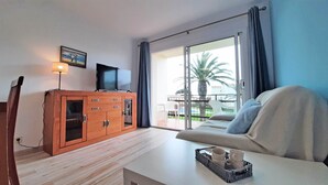 Ein Zimmer mit einer Couch, einem Fernseher auf einer Holzkommode, einem Tisch mit Tabletts voller Tassen und Tellern sowie einer Glastür, die zu einem Außenbereich mit einer Palme führt.