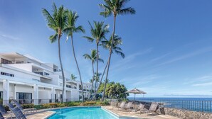 Un área junto a la piscina con sillones de descanso, una piscina, palmeras y vista al océano.