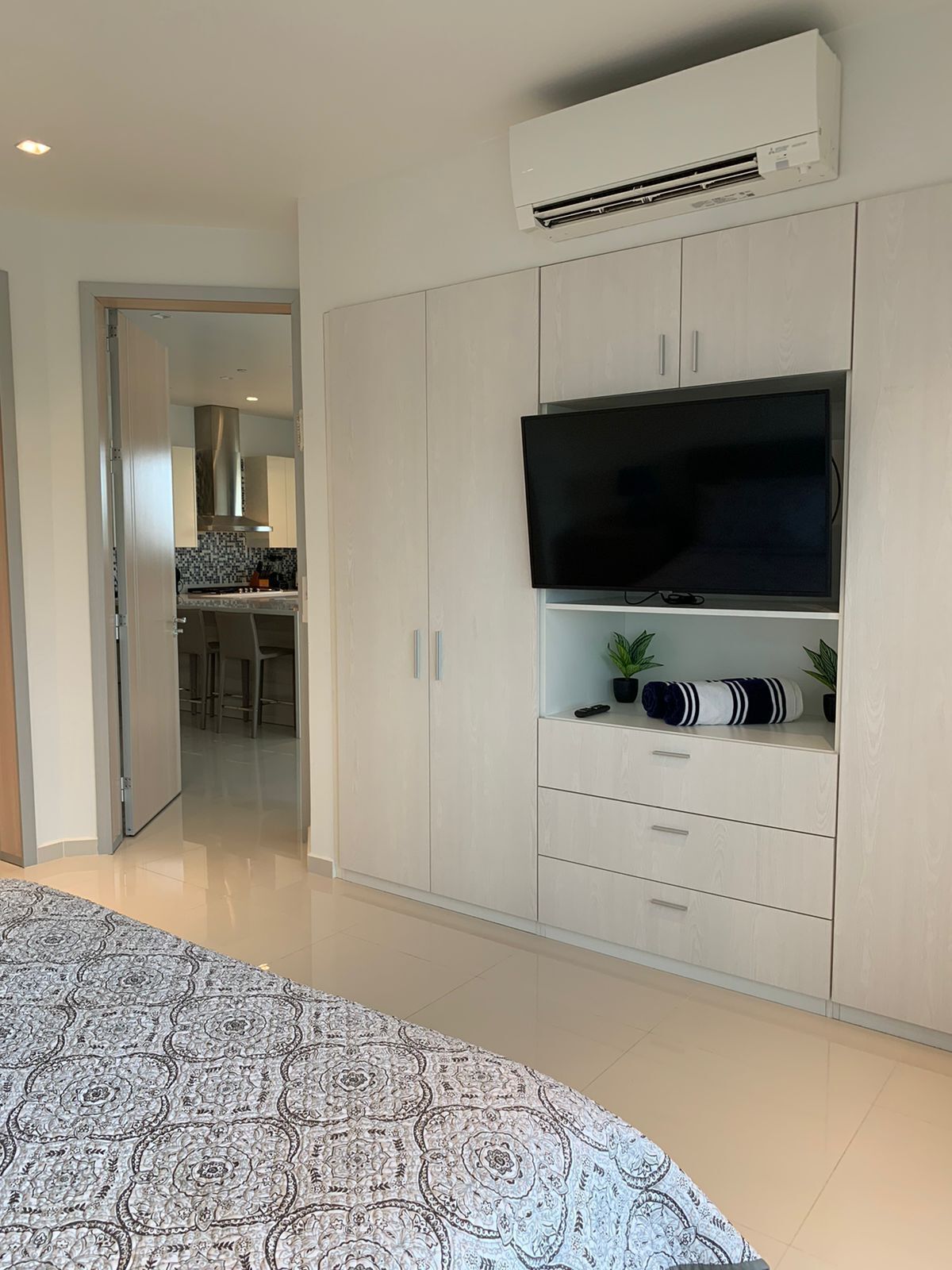 A modern bedroom with a large bed, a TV mounted on a white wardrobe, and an air conditioning unit above the TV.