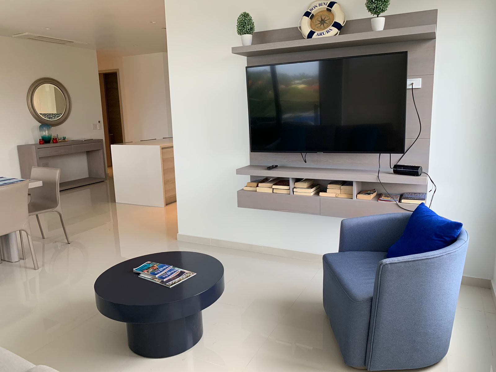 Een moderne woonkamer met een flatscreen televisie aan de wand, een grijze bank met een blauw kussen, een ronde zwarte salontafel en een klein boekenrek onder de televisie.