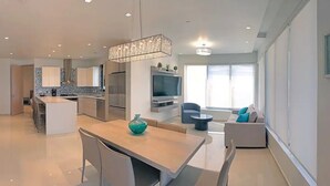 Una cucina moderna con isola centrale, un'area pranzo con tavolo e sedie, un soggiorno con divano e televisore, e un'ampia finestra che permette l'ingresso di luce naturale.