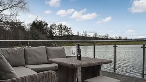 Ein Sitzbereich im Freien mit Korbmöbeln, einem Tisch und Kissen, mit Blick auf ein Gewässer und Bäume.