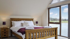 Ein Schlafzimmer mit einem hölzernen Bett, zwei Nachttischen, einem Beistelltisch und einem großen Fenster mit Blick auf einen See und Bäume.