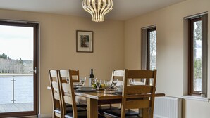 Ein Essbereich mit einem Tisch für vier Personen, Holzsesseln, einer Chandelüstere und einem Fenster mit Blick auf ein Gewässer.
