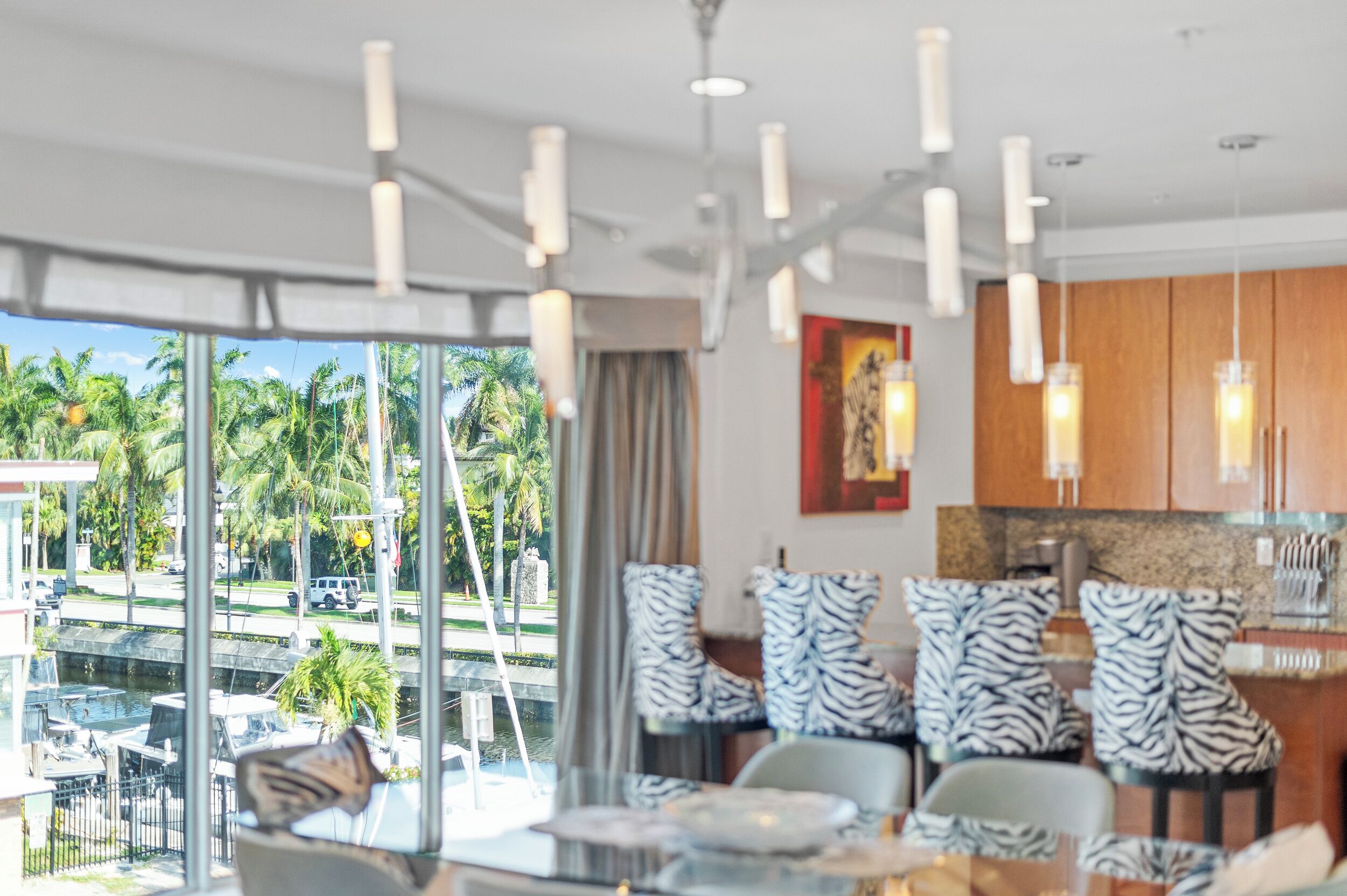 Una habitación de hotel moderna con un ventanal que ofrece vistas a palmeras y un puerto deportivo. Hay sillas con estampado de cebra y una mesa de comedor.