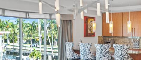 Una habitación de hotel moderna con un ventanal que ofrece vistas a palmeras y un puerto deportivo. Hay sillas con estampado de cebra y una mesa de comedor.