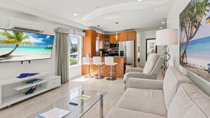 A modern living room with a TV, sofa, and a glass coffee table. There is a kitchen area with a bar and seating. - Isle of Venice (Fort Lauderdale)