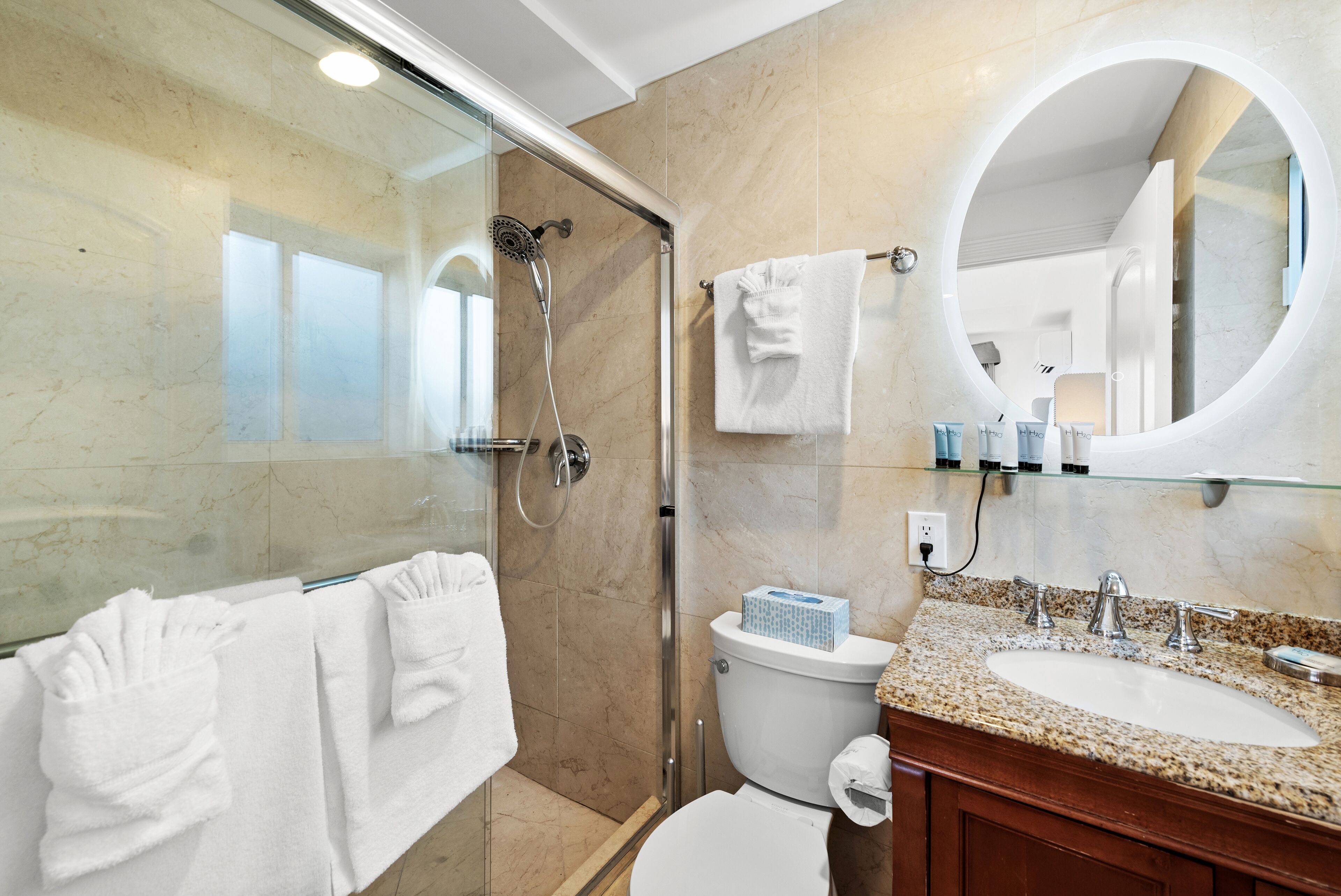 A modern bathroom with a glass-enclosed shower, a white toilet, a sink with a granite countertop, and a round mirror.