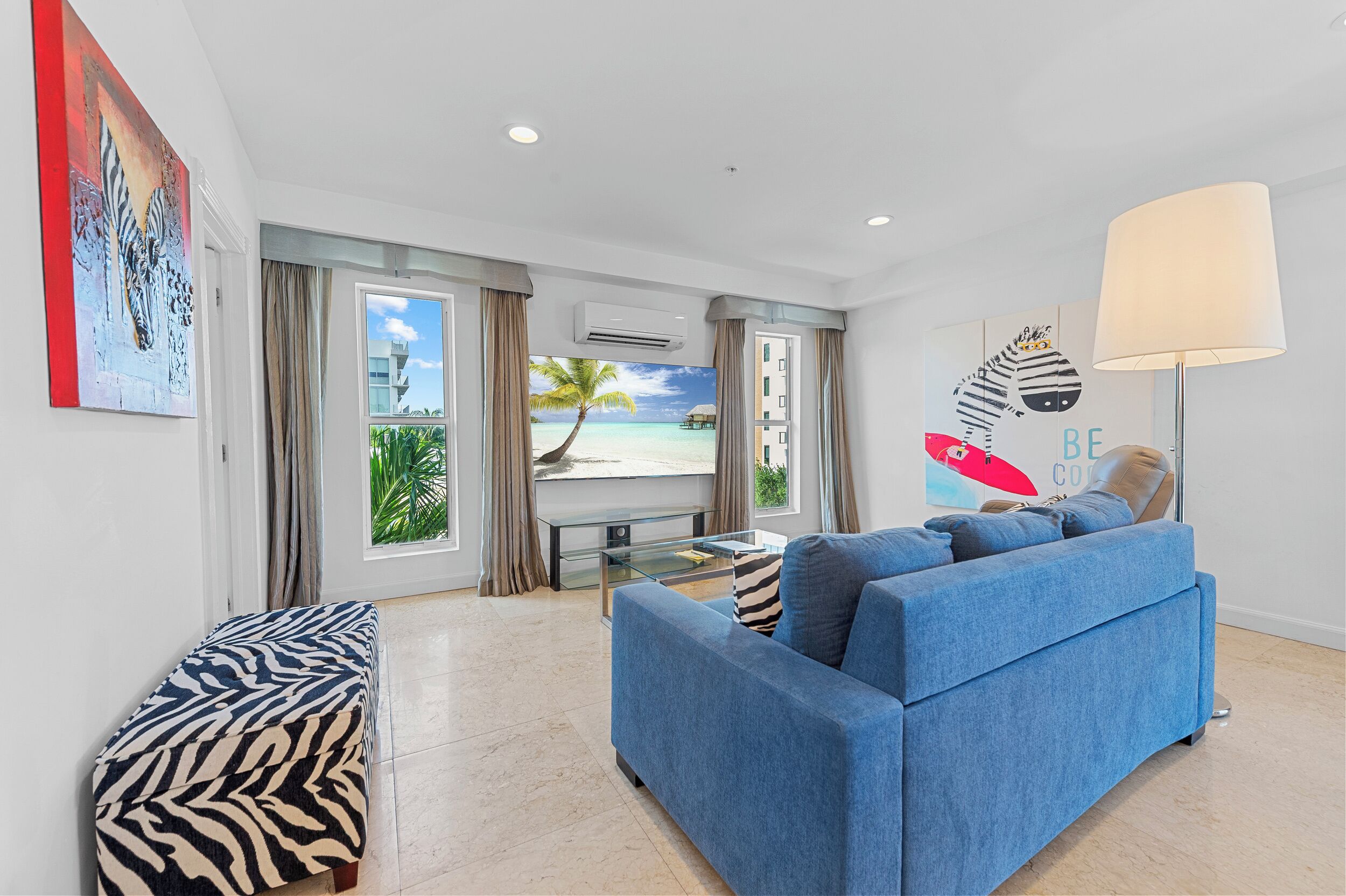 Un salon moderne avec un canapé bleu, un banc à motifs zébrés et une grande fenêtre donnant sur une plage.