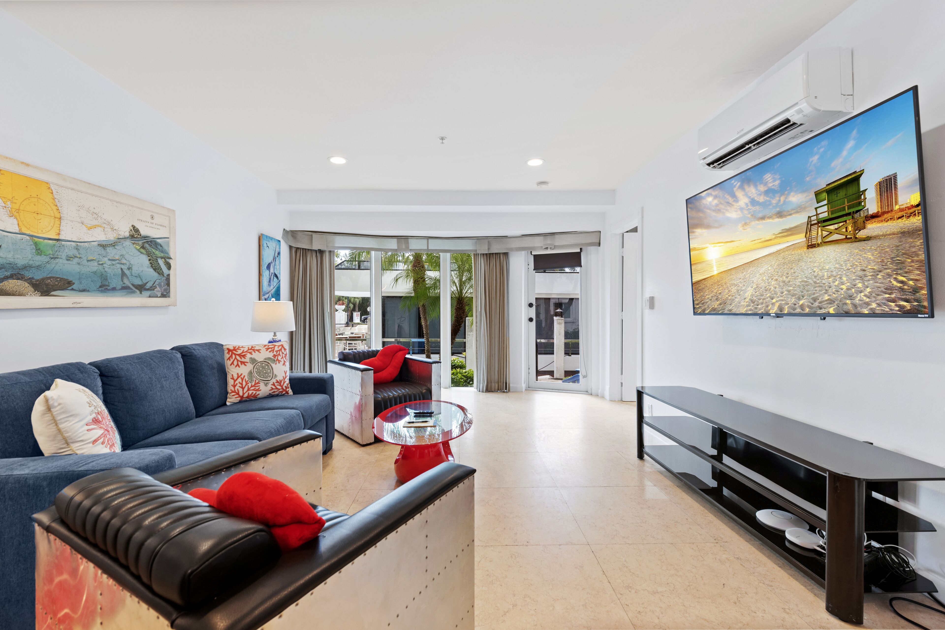 Una sala de estar moderna con un sofá azul, un cojín rojo, una mesa de centro de vidrio y un televisor plano grande montado en la pared.