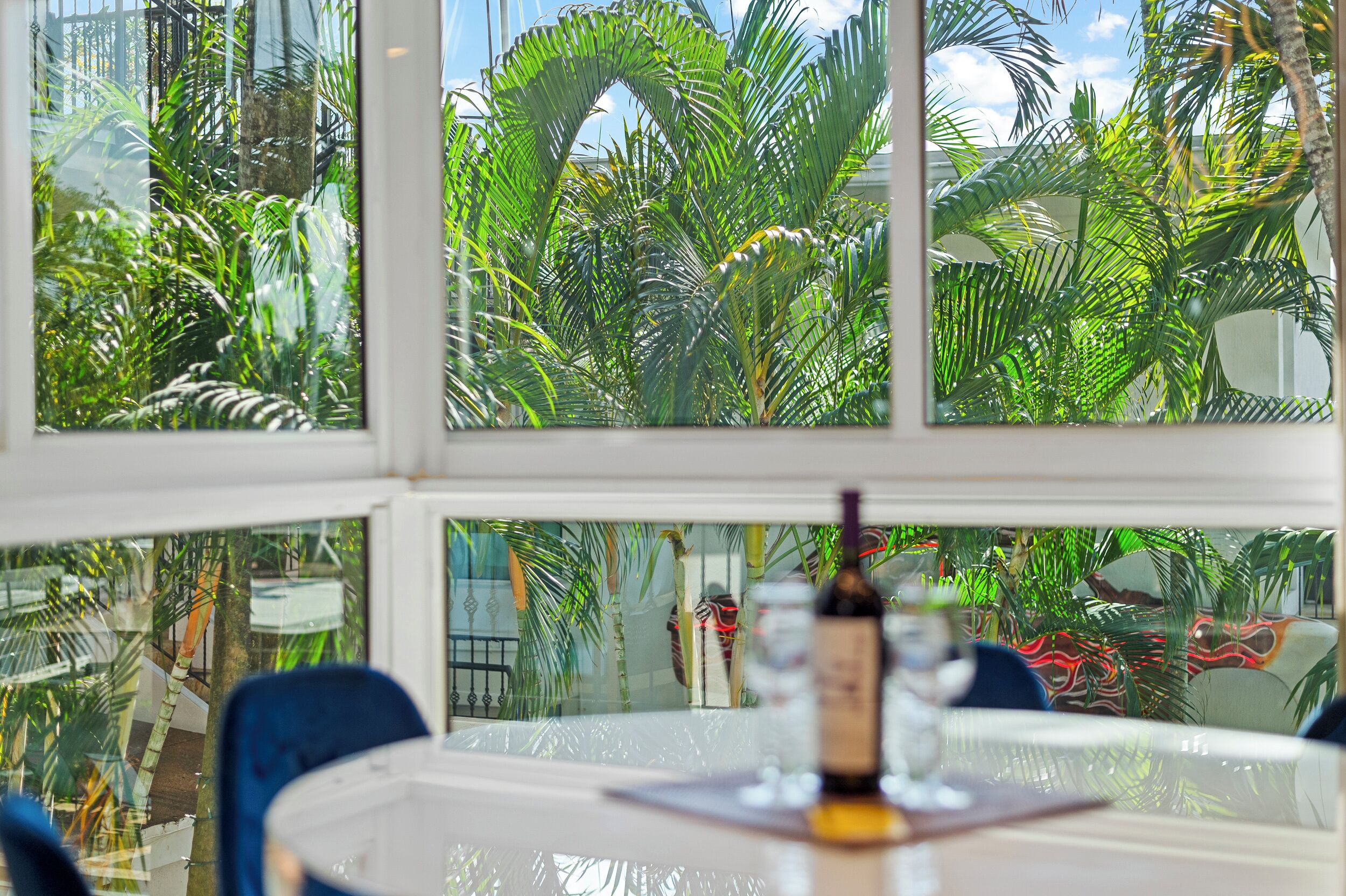A dining area with a table and chairs, a bottle of wine, and a view of lush tropical plants through large windows.
