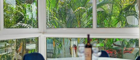 A dining area with a table and chairs, a bottle of wine, and a view of lush tropical plants through large windows.