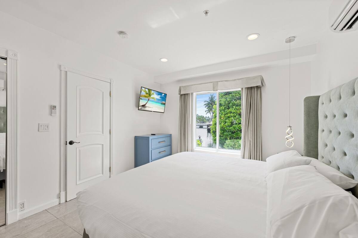 Quarto moderno com uma cama grande, uma cômoda azul, um quadro de uma praia e vista para o verde pela janela.