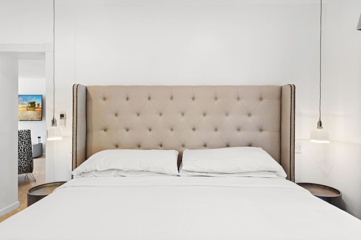 Cama bem arrumada com cabeceira estofada, roupa de cama branca e mesa de cabeceira com abajur.