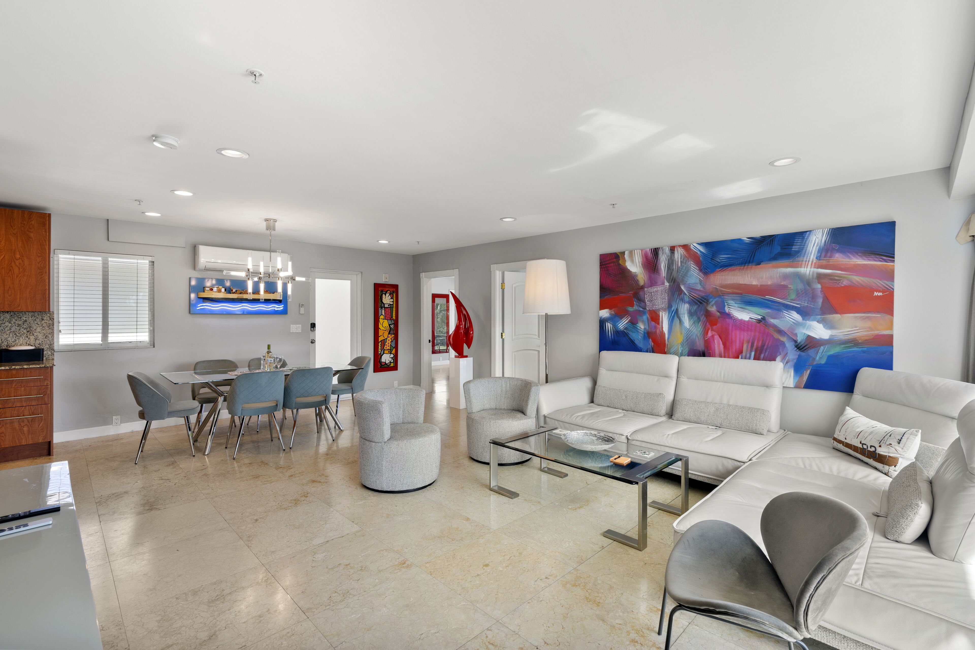 A modern living room with a white sofa, grey armchairs, a glass coffee table, and a dining area with a white table and grey chairs.