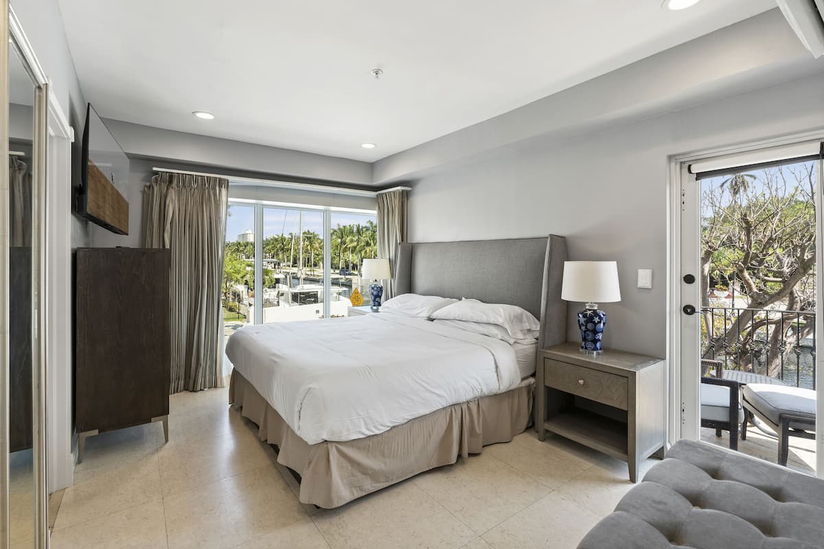 Quarto de hotel moderno com uma cama grande, uma mesa de cabeceira com abajur, um espaço de estar com mesa e cadeiras, e uma varanda com vista para palmeiras.