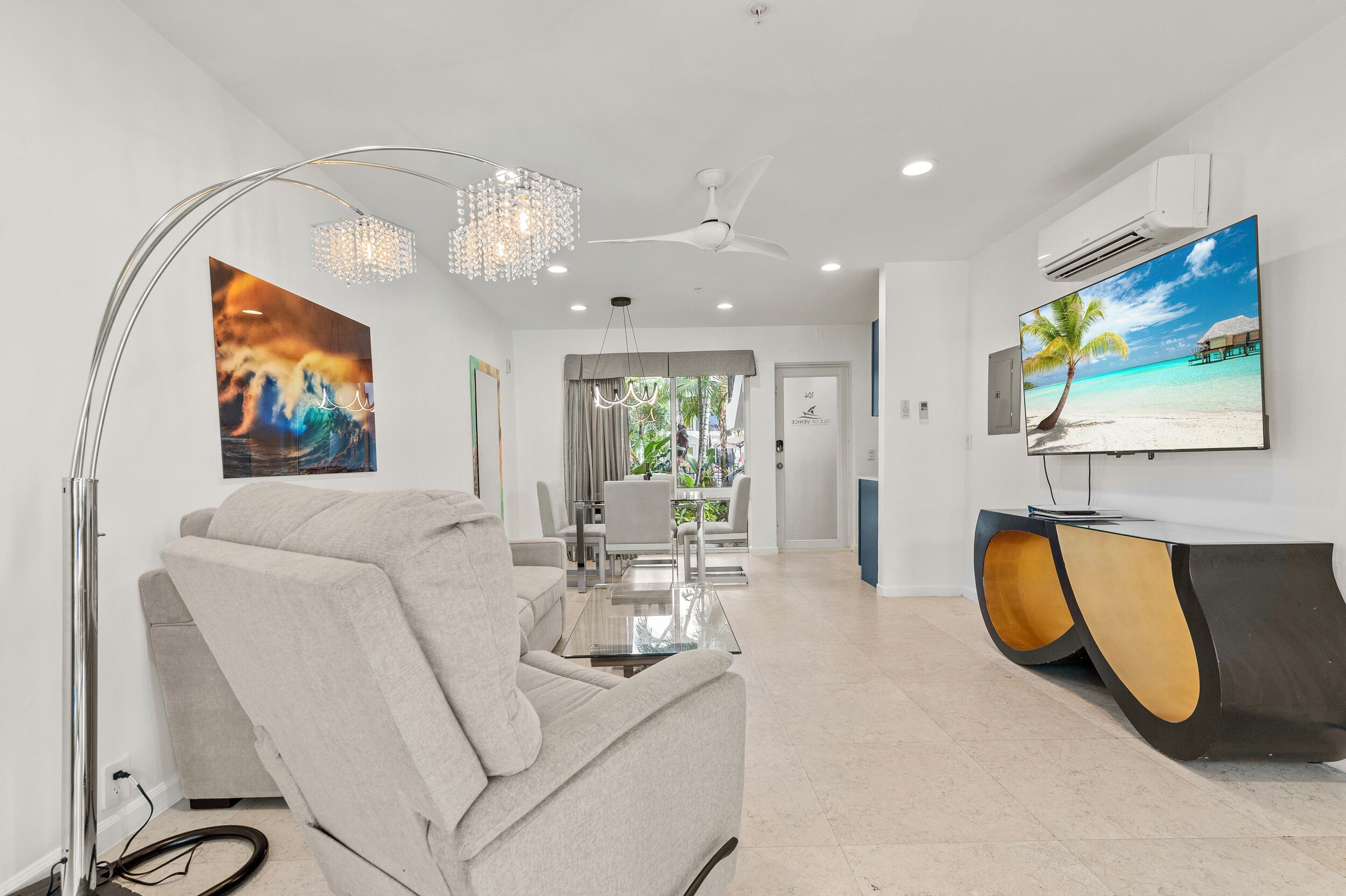 A modern living room with a reclining chair, a glass coffee table, a chandelier, a ceiling fan, and a large flat-screen TV displaying a beach scene.