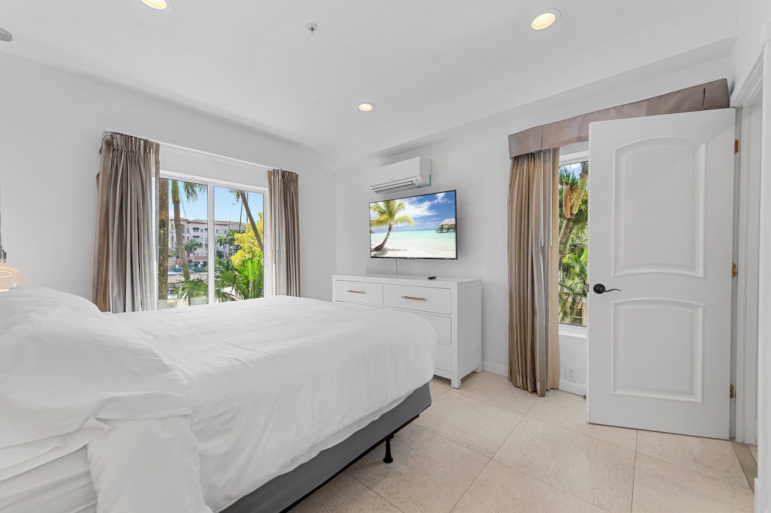 A hotel room with a large bed, a TV, a white dresser, and a door leading to a balcony with a view of palm trees and a beach.