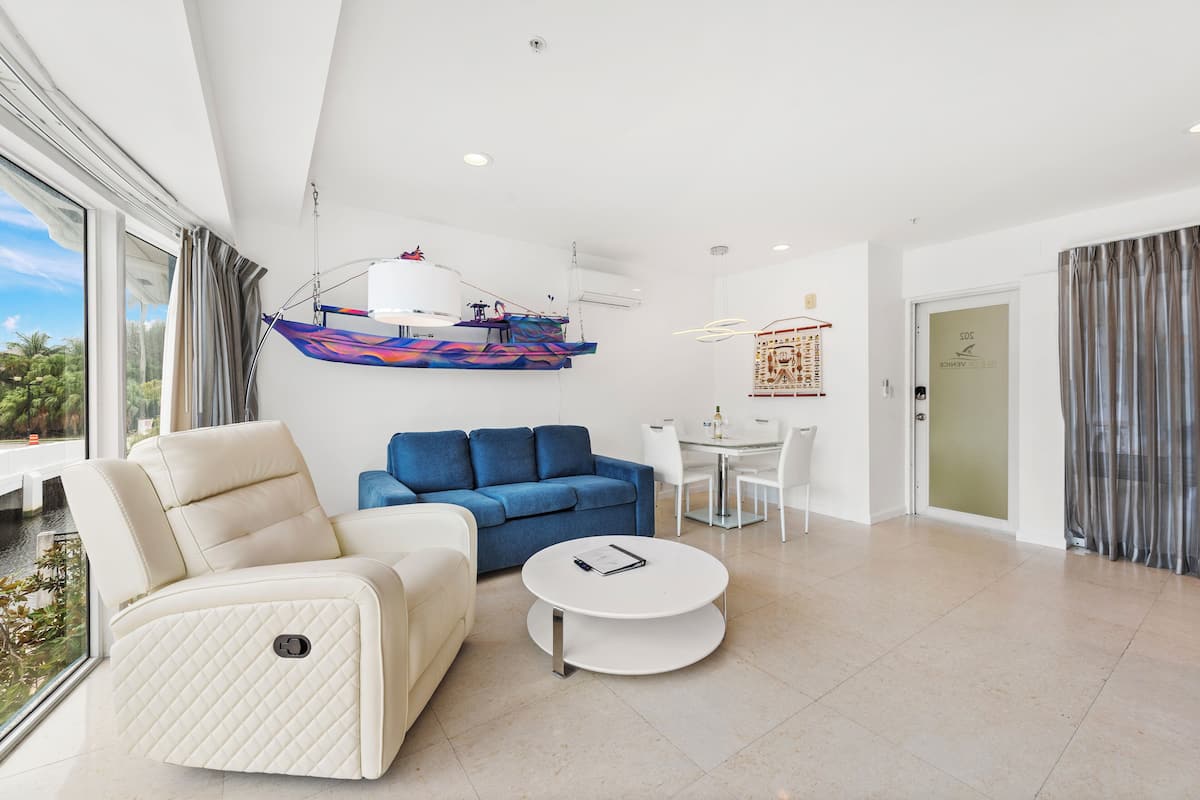 Uma sala de estar moderna com um sofá branco, um sofá azul, uma poltrona branca, uma mesa de centro redonda e uma área de jantar com mesa e cadeiras de vidro.