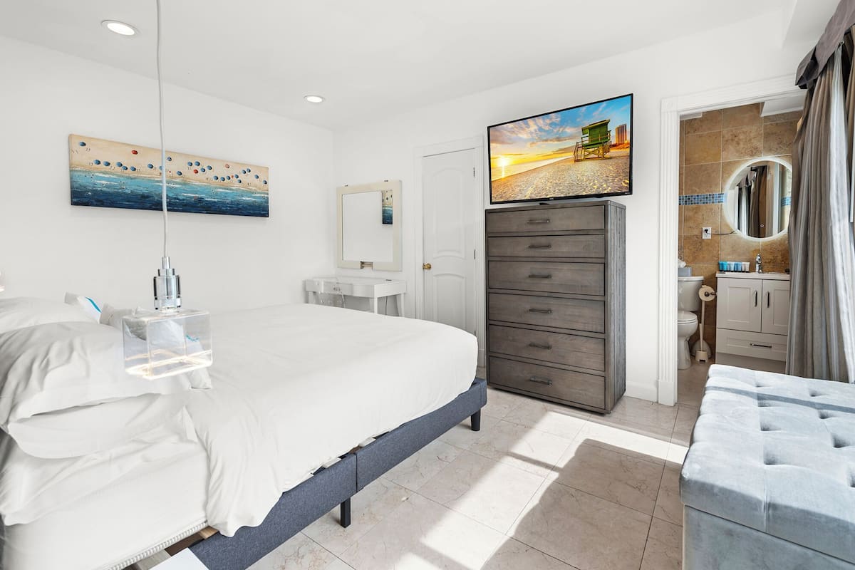 Quarto de hotel moderno com uma cama grande, televisão de tela plana, banheiro com vaso sanitário e pia, e vista para a praia.