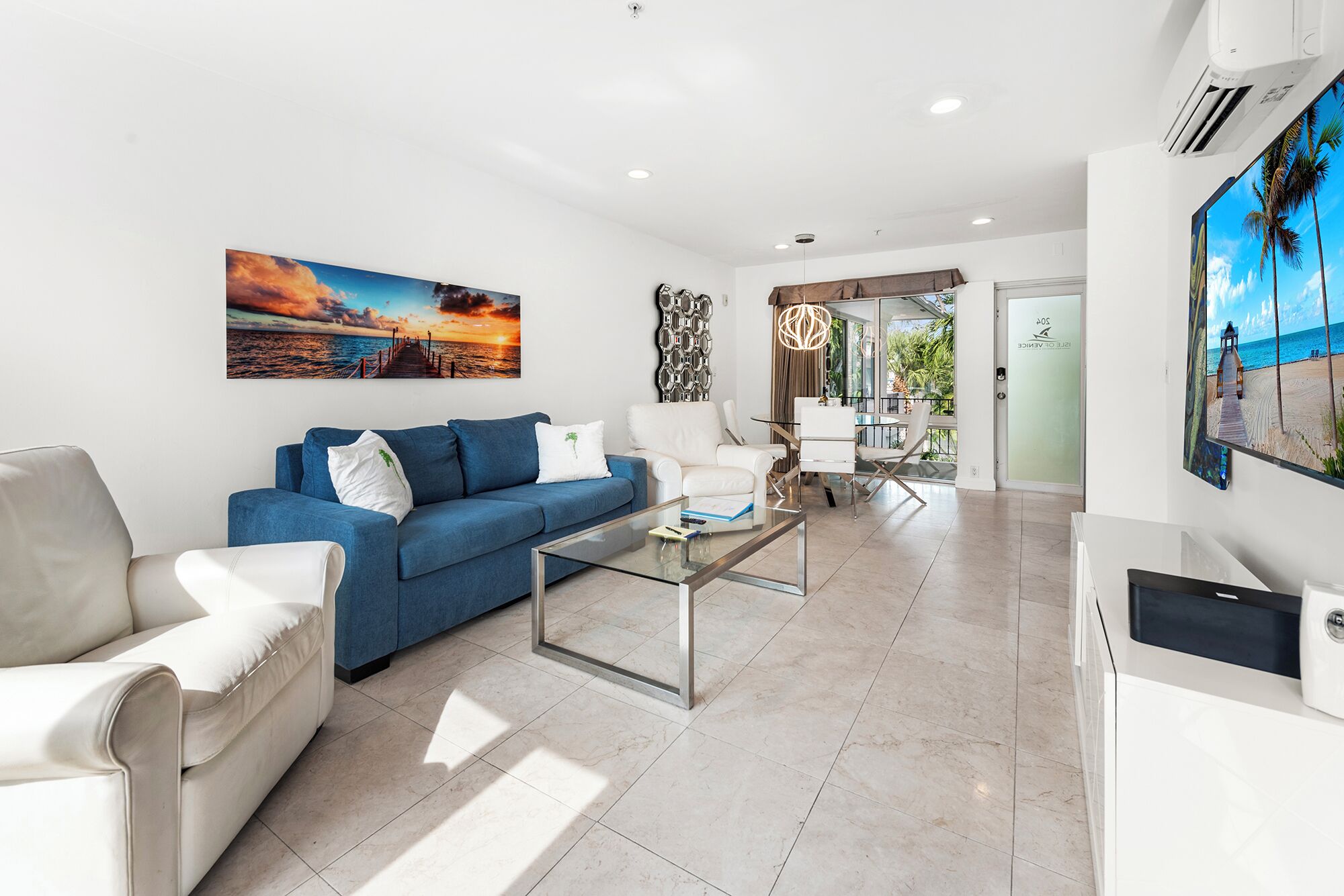 Un salon moderne avec un canapé bleu, des fauteuils blancs et une table basse en verre. Un tableau représentant un coucher de soleil est accroché au mur, et une porte donne accès à un balcon avec vue sur la plage.