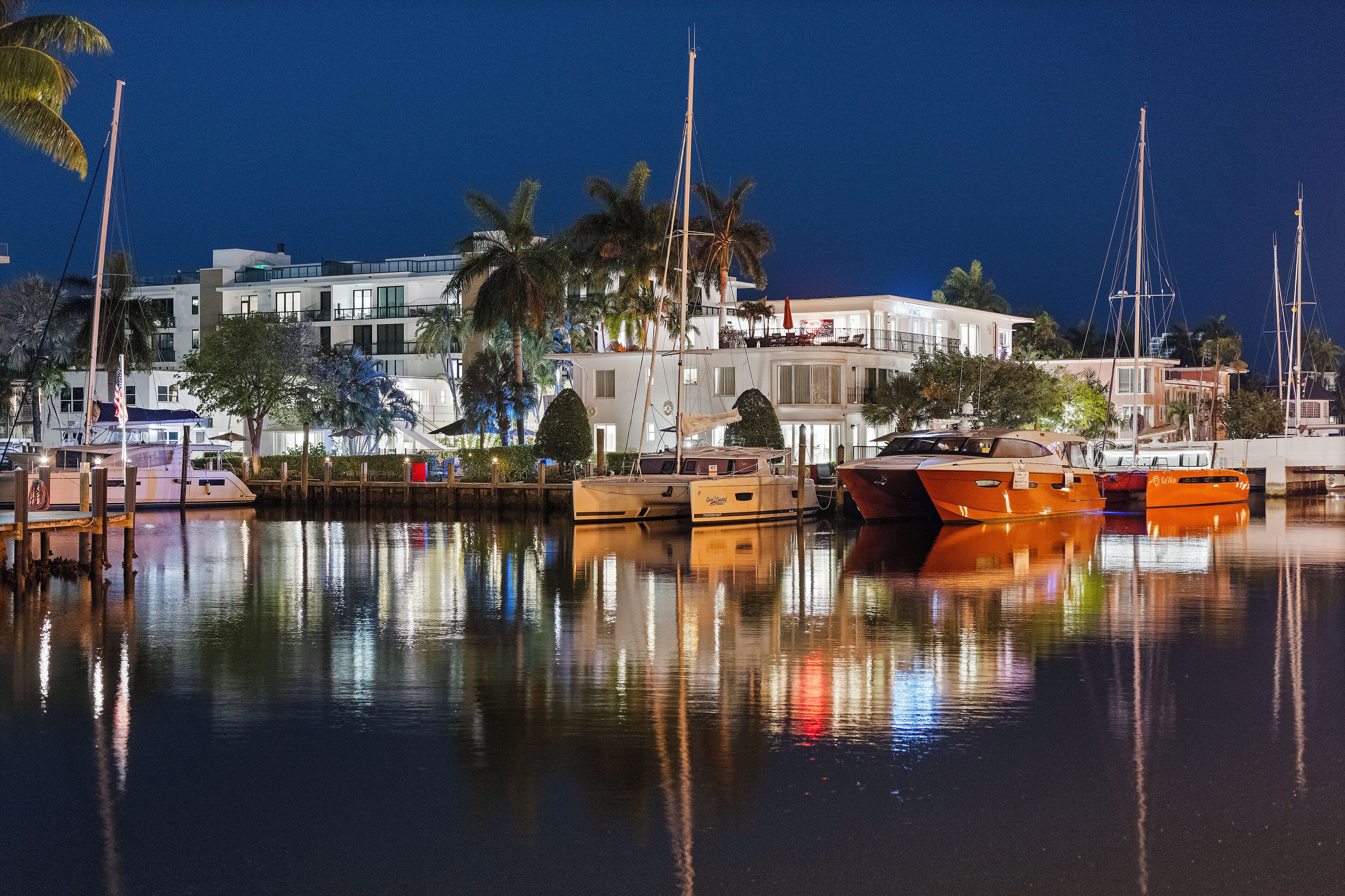 A marina with boats docked, a building with balconies, and palm trees.