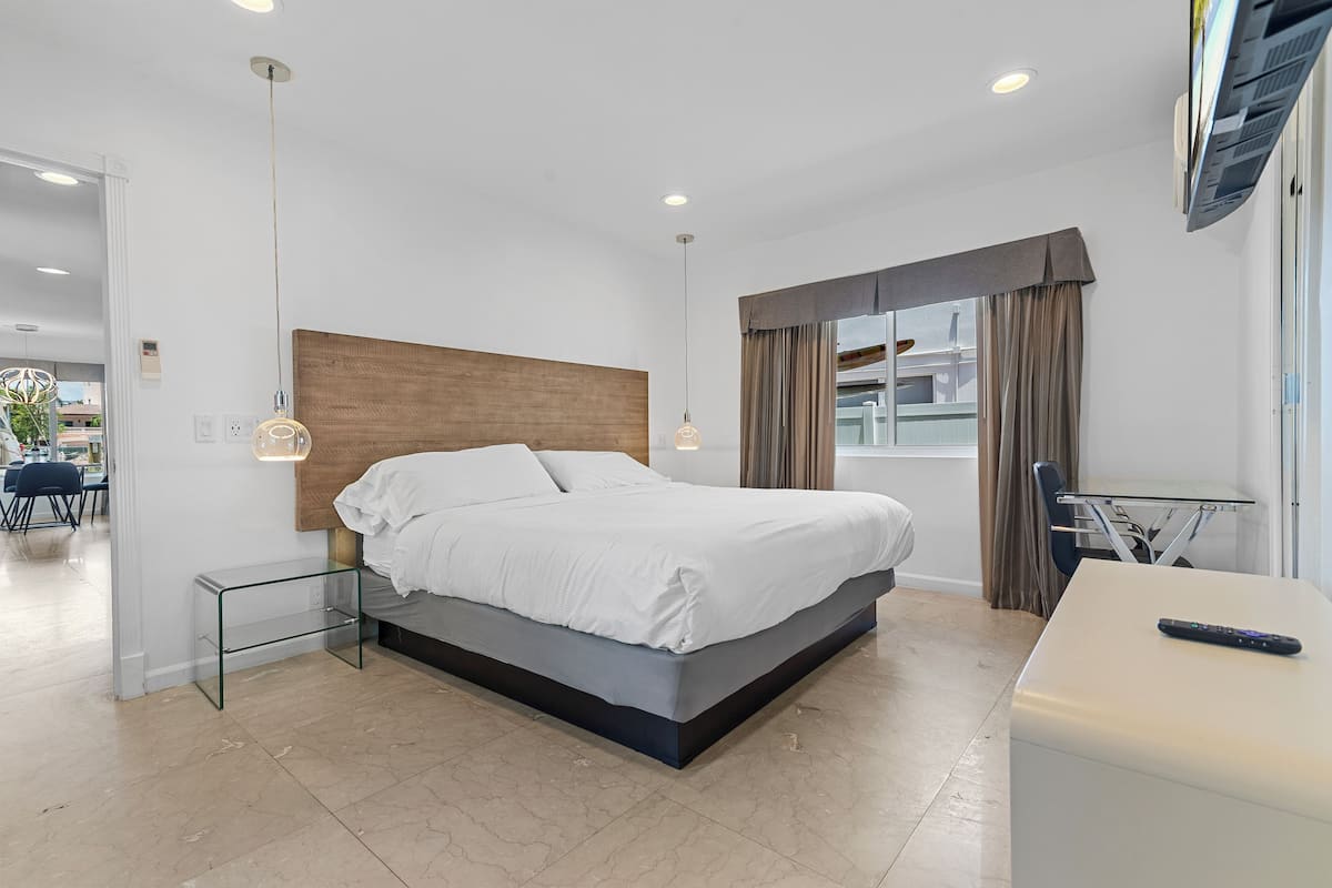 Quarto de hotel moderno com uma cama grande, mesa de cabeceira de vidro, pequena escrivaninha com cadeira e televisão fixada na parede.