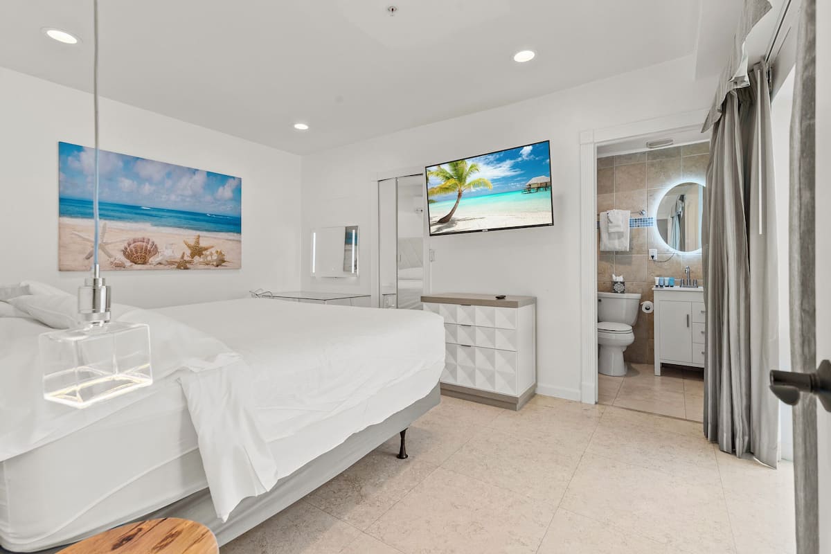 Quarto moderno com cama, mesa de cabeceira com abajur, televisão e banheiro visível por uma porta aberta.