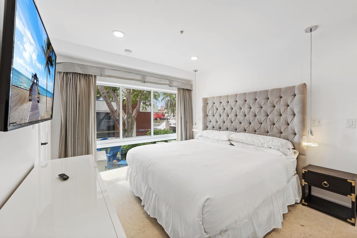 Um quarto moderno com uma cama grande, uma TV na parede e vista para a praia pela janela.