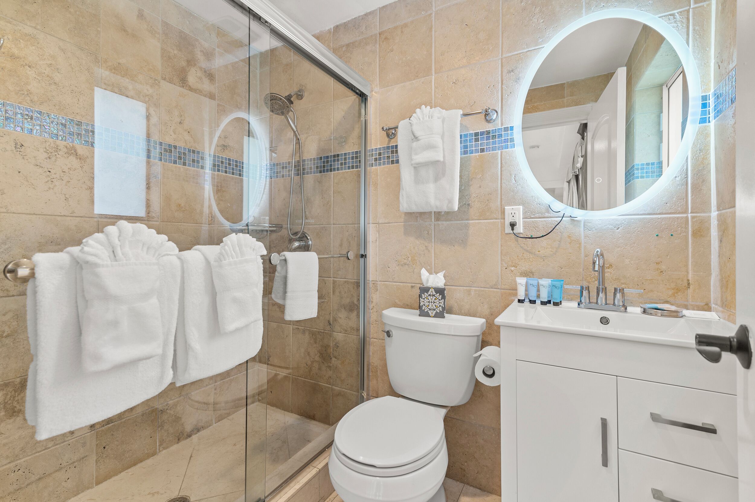 Un baño moderno con cabina de ducha de vidrio, inodoro, lavamanos con artículos de aseo y espejo.