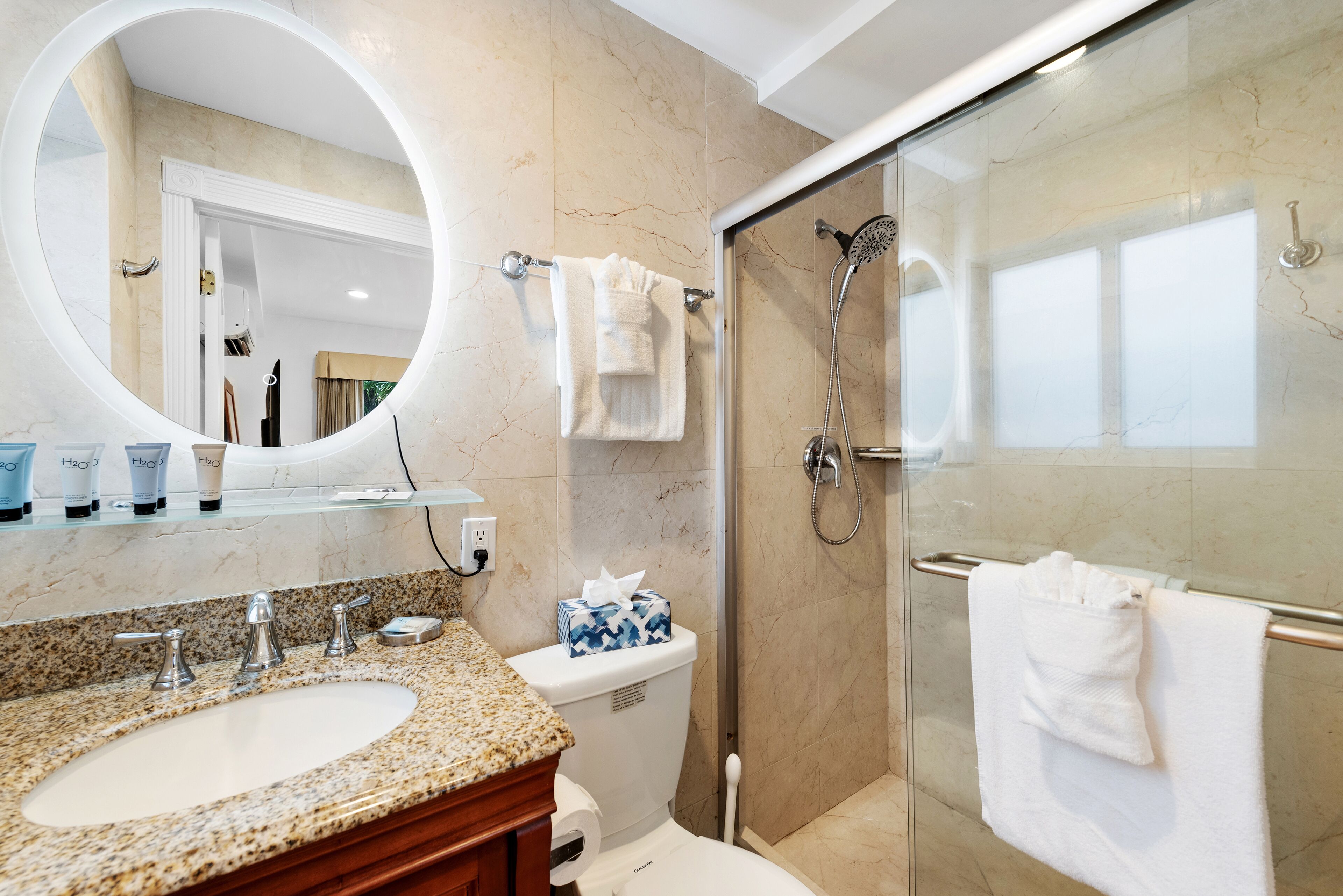 A modern bathroom with a granite countertop sink, a toilet, a glass-enclosed shower, and a large mirror with a circular frame.