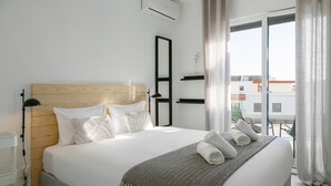 A modern hotel room with a large bed, a wooden headboard, a bedside table with a lamp, and a view of the city through large windows. - Sunny Penthouse - Modern & Premium Apartment (Peniche)