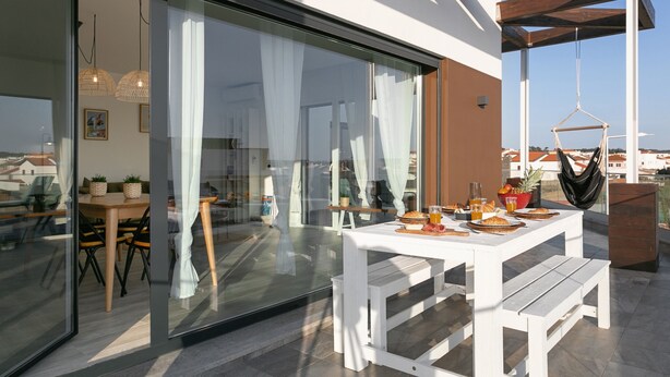 A modern outdoor dining area with a white table and benches, a hammock, and a glass door leading to an interior space. - Sunny Penthouse - Modern & Premium Apartment (Peniche)