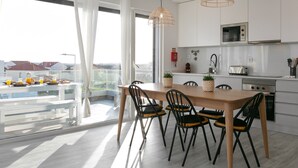 A modern kitchen with white cabinets, a wooden dining table, and black chairs. There is a large window with a view of buildings outside. - Sunny Penthouse - Modern & Premium Apartment (Peniche)