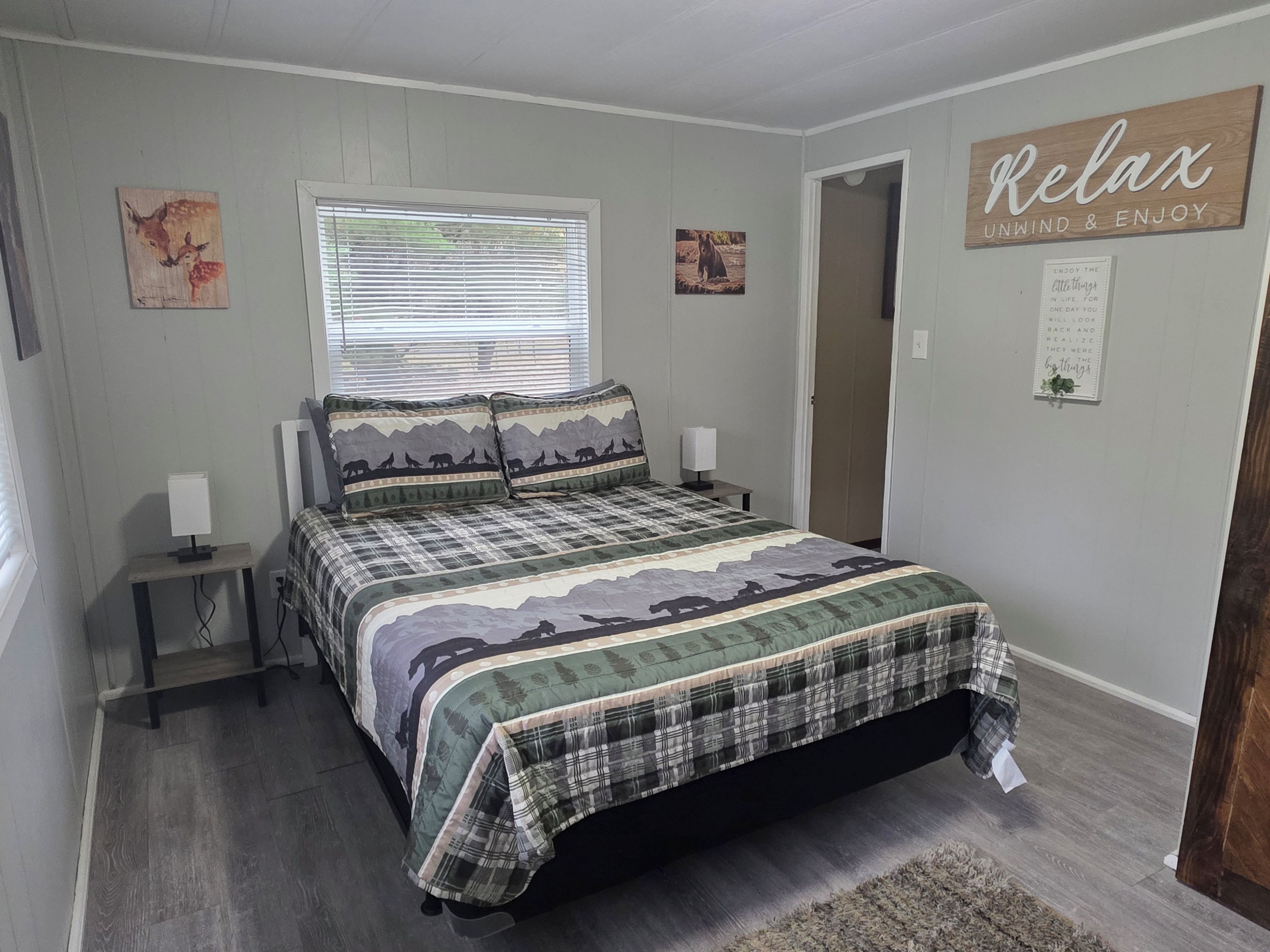 A bedroom with a bed, a bedside table, a lamp, a window with blinds, and a wall with a 'Relax' sign.