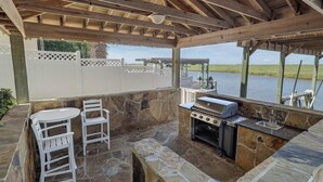 An outdoor patio with stone flooring, a grill, white chairs, and a view of a body of water. - Coastal Condo w/ Pool, Balconies, Sundeck & Boat Slip - Near Beach (Corpus Christi)