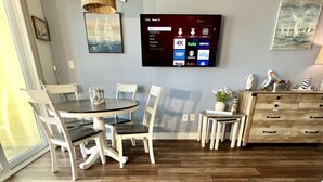 A modern living room with a wooden dining table, chairs, a TV mounted on the wall, and a potted plant on the floor.