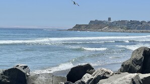 A coastal scene with waves, rocks, and a distant building complex.