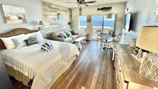 A spacious bedroom with a large bed, a sitting area with a sofa and chairs, a dining table, and a balcony with a view of the ocean.