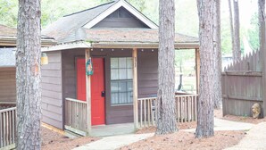 A small cabin with a red door, surrounded by trees, and a wooden deck. - 1 rm cabin, shared bth house, at Kick Back Ranch. (Ramer)