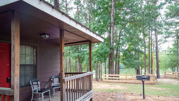 A wooden cabin with a porch, two chairs, and a red door. - 1 rm cabin, shared bth house, at Kick Back Ranch. (Ramer)